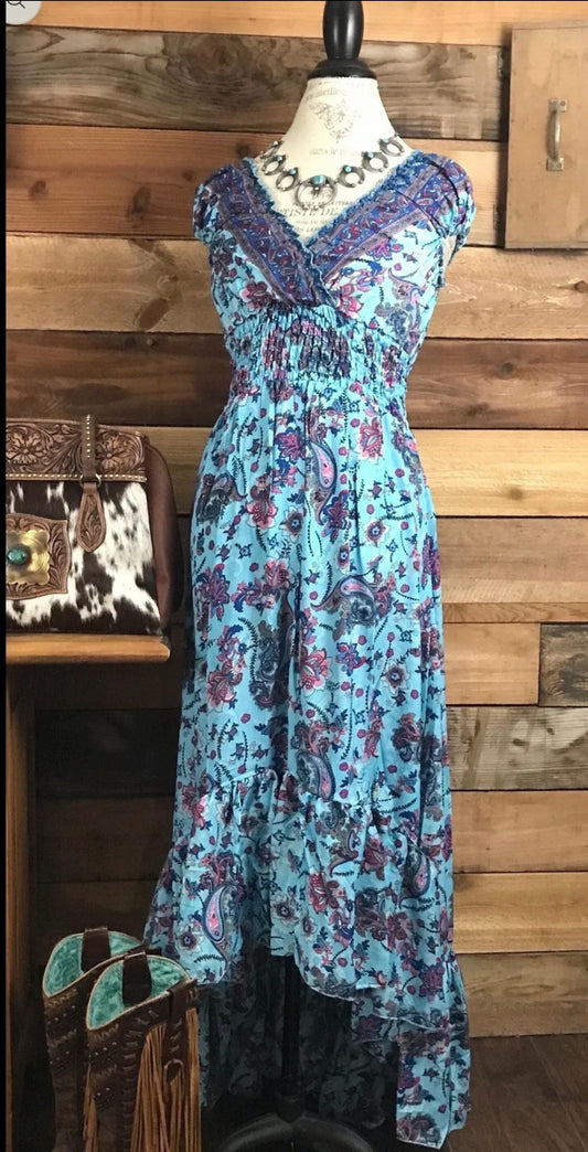 May include: A blue and purple paisley print maxi dress with a high-low hemline. The dress has a v-neckline and short sleeves. The dress is on a mannequin and has a silver necklace with turquoise stones.